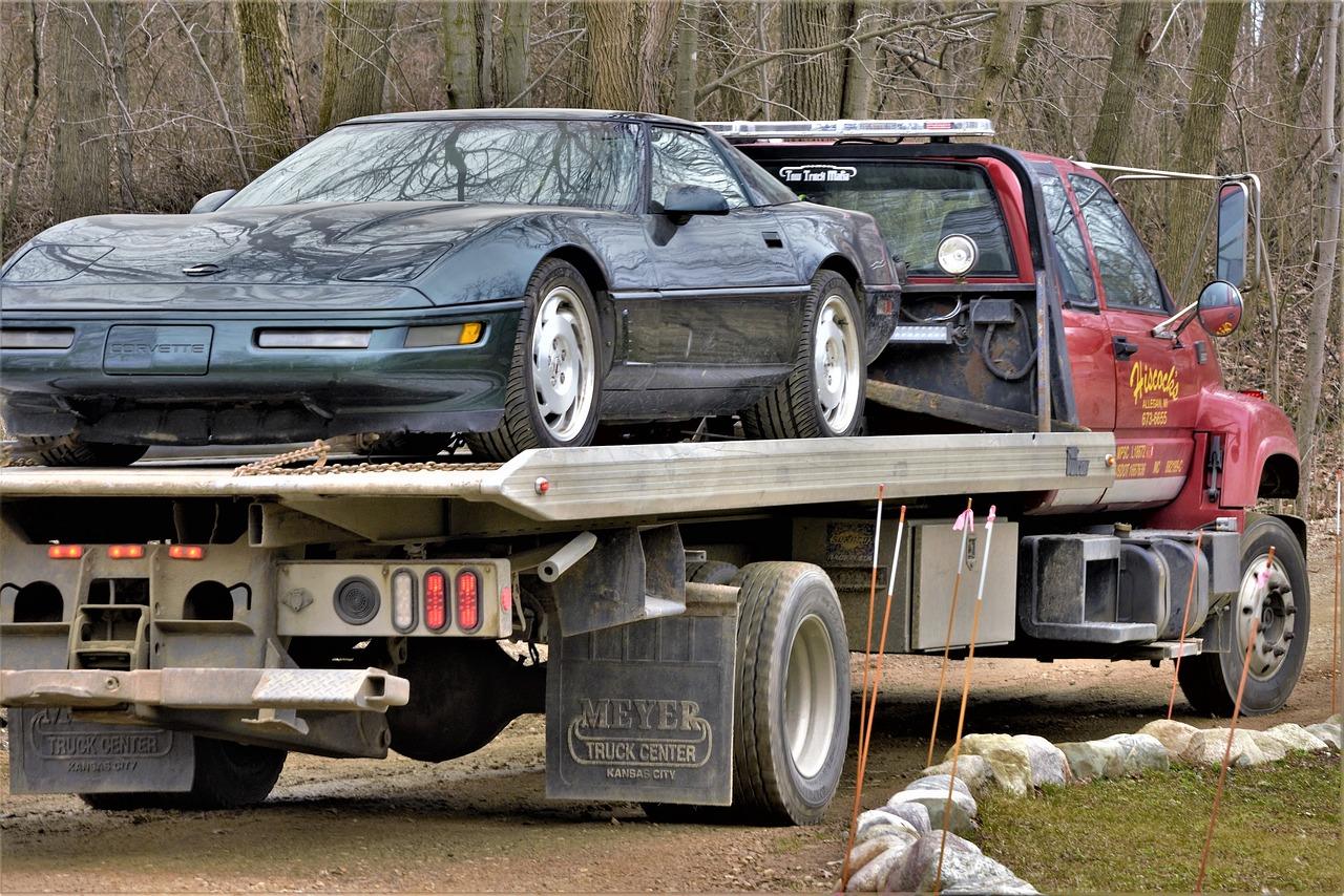 A red flatbed tow truck carrying a dark green Corvette on its bed