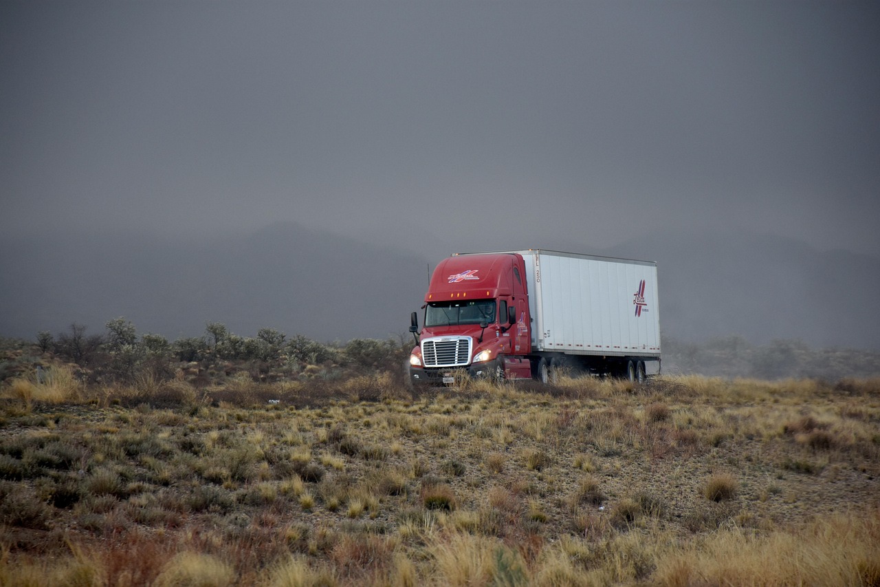 A red semi-truck with a white trailer on a grassland road under a stormy sky